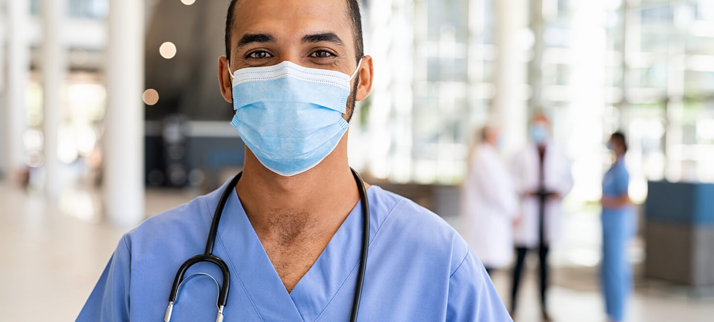 A doctor wearing scrubs, a stethoscope, and a medical mask appears to be smiling at the camera.