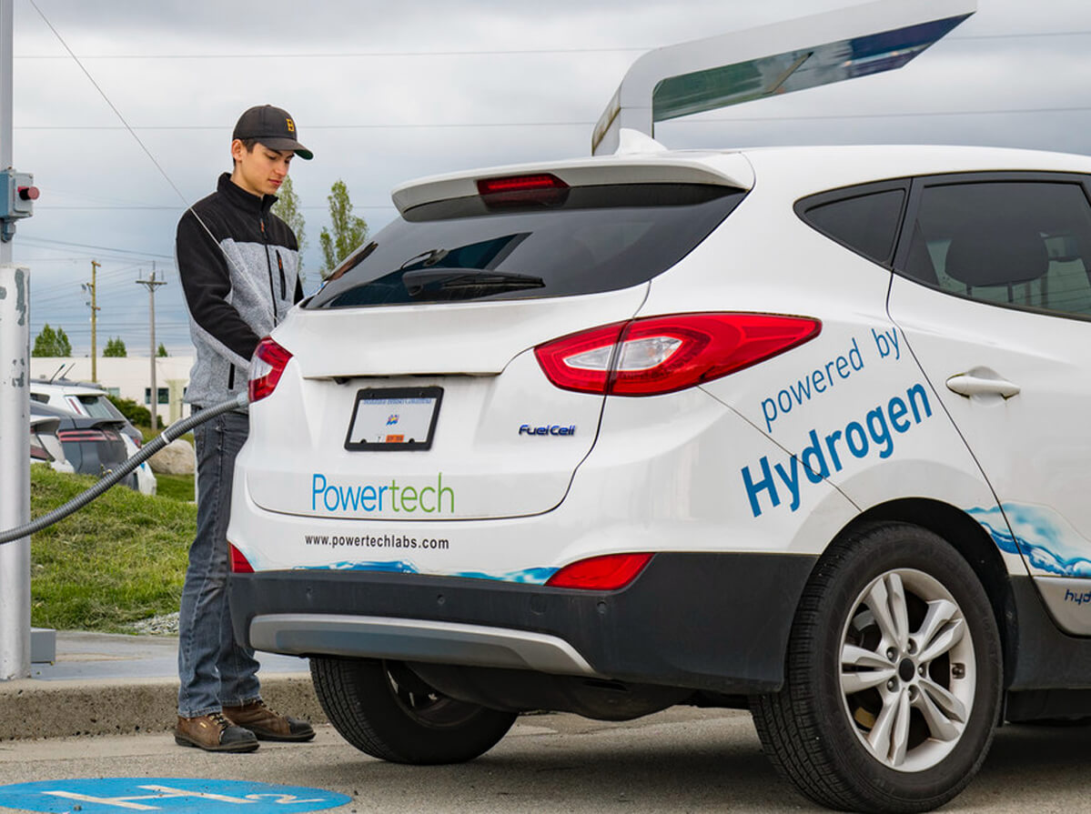 A man fuelling a Fuel Cell SUV. The vehicle is white with the words "Power tech" and "powered by Hydrogen" along its body.