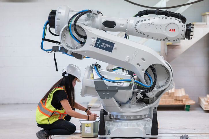 A worker wearing a hard hat, mask, and hi-vis vest kneels to work on a large pneumatic machine.