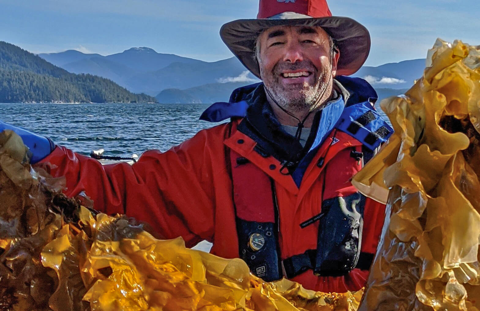 A white man wearing red waterproof gear holds up a large piece of kelp with two hands, smiling at the camera.