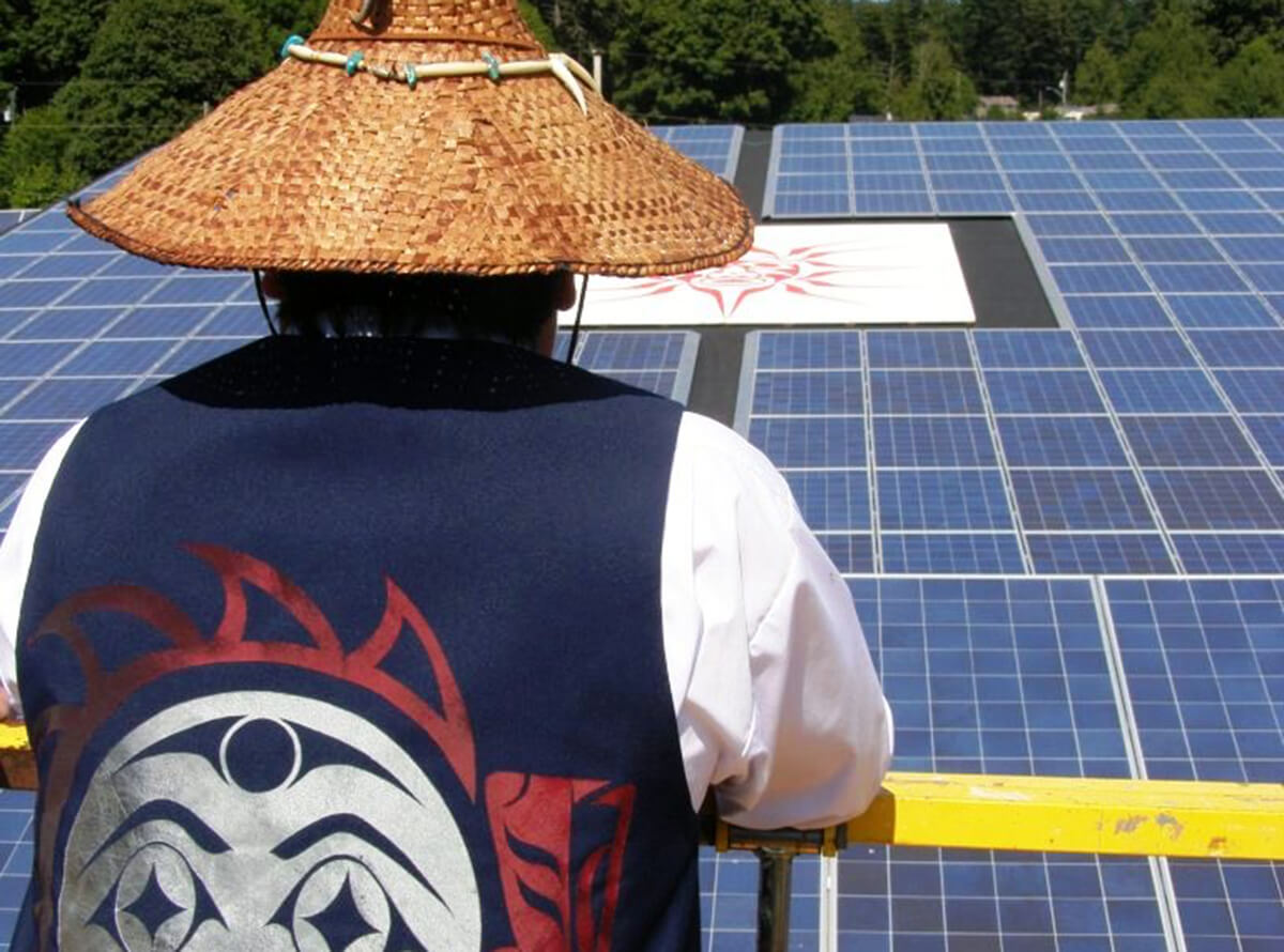An Indigenous person wearing a vest with traditional art and a woven hat looks out at a large solar panel with traditional art placed in the centre.