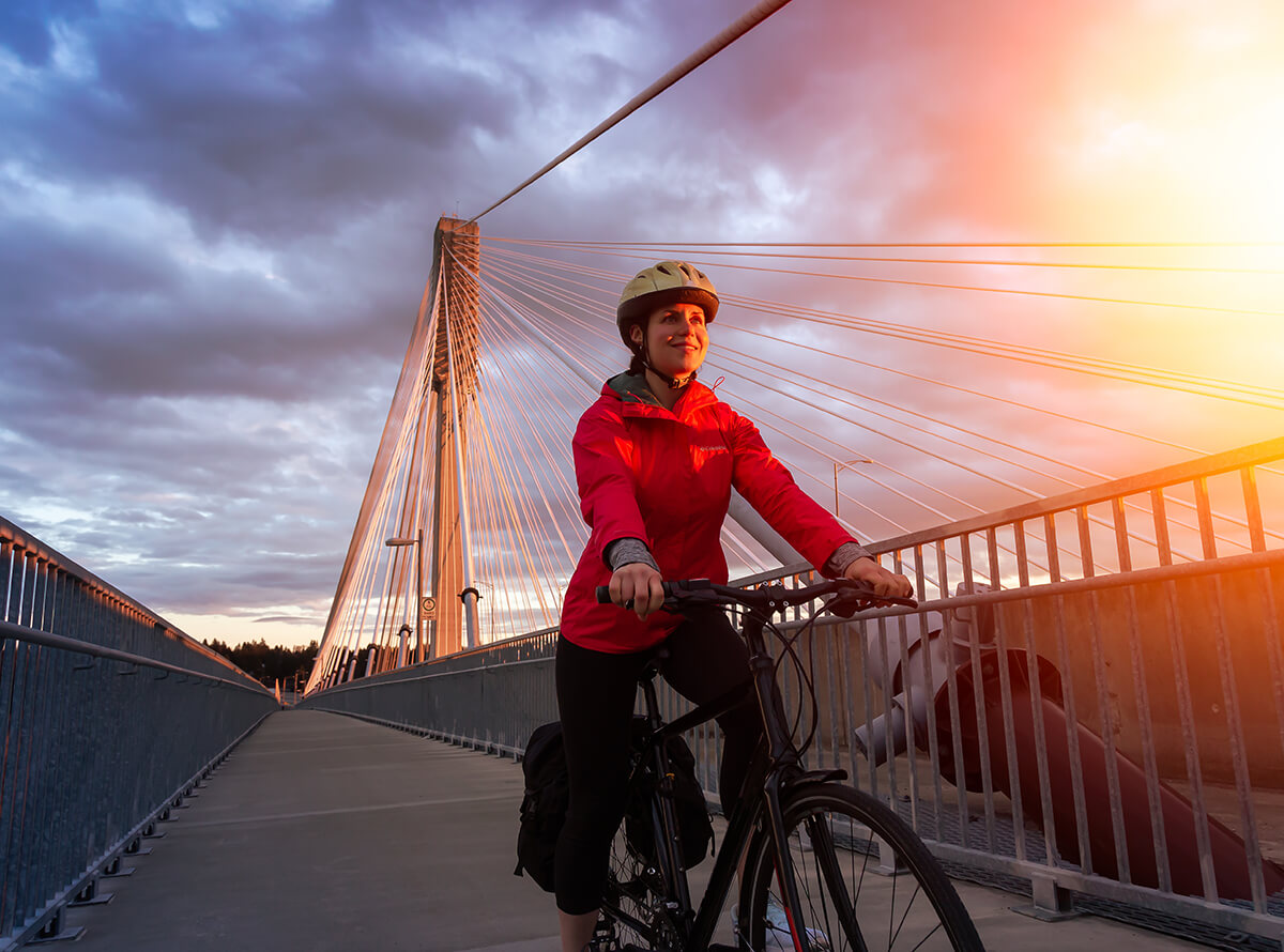 A cyclist wearing a bright red jacket poses on the Alex Fraser bridge around sunset.
