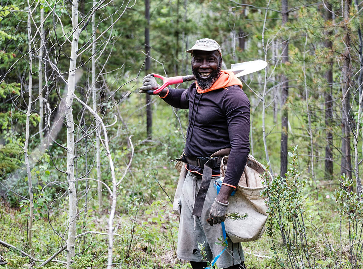A Black man holds a spade over his shoulder with a large sack hanging on his hip. He is working in a forested area with young trees.