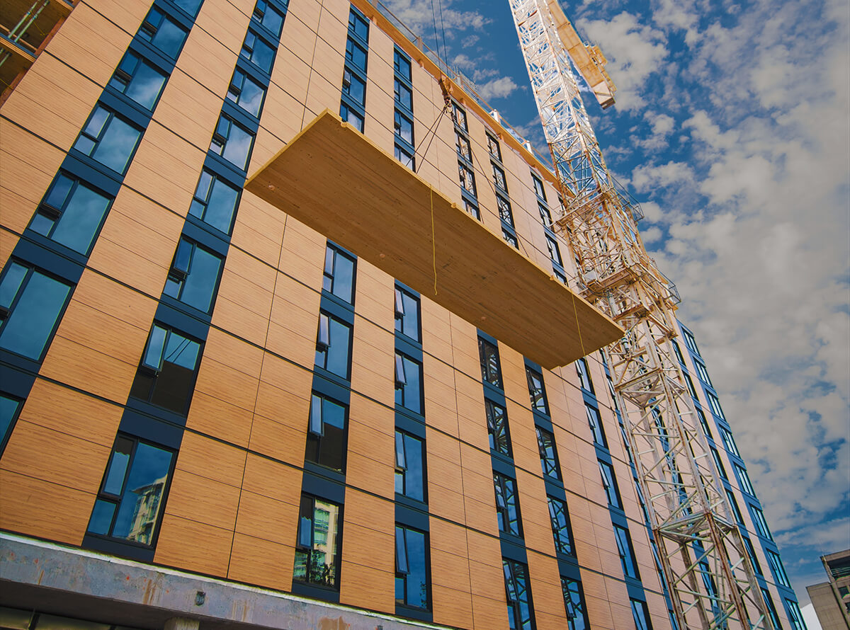A large panel of wood is seen from below as it is lifted by a crane to the top of a tall wood-panelled building being constructed.