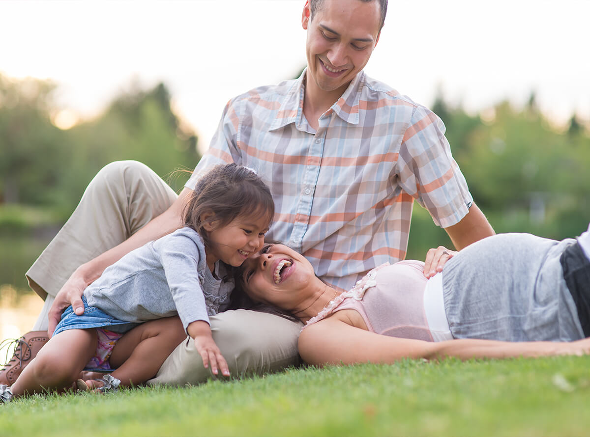 A family sit and lay on the grass together, laughing. The mother has her head on the father's lap while they smile at their toddler climbing over them. The mother appears pregnant.