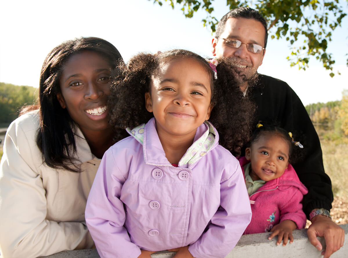 A young biracial family with two small girls smile at the camera while under a tree outside.
