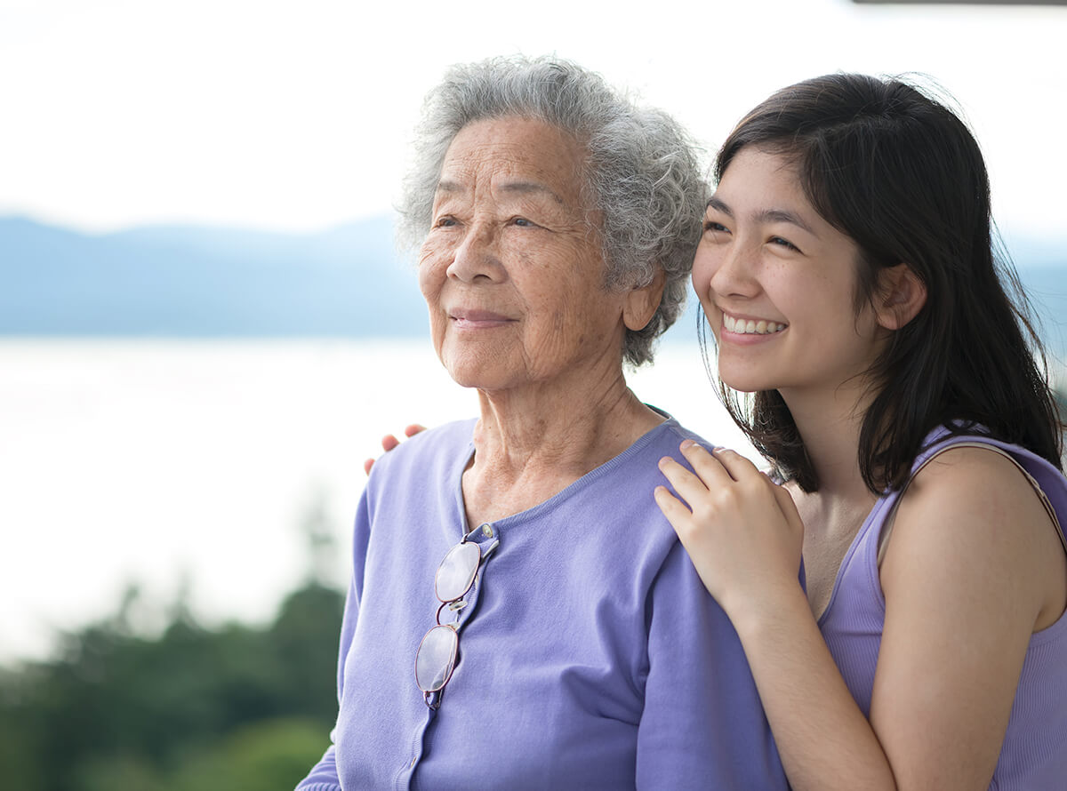 A young Asian woman and her grandmother both wear lavender shirts outside as they smile and look out into the distance together.