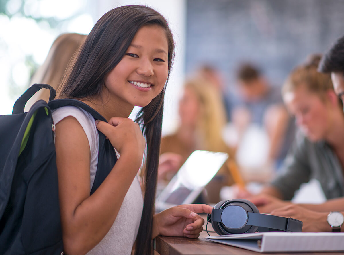 A young teen smiles at the camera with her backpack on her shoulder while standing at a raised table next to other people.