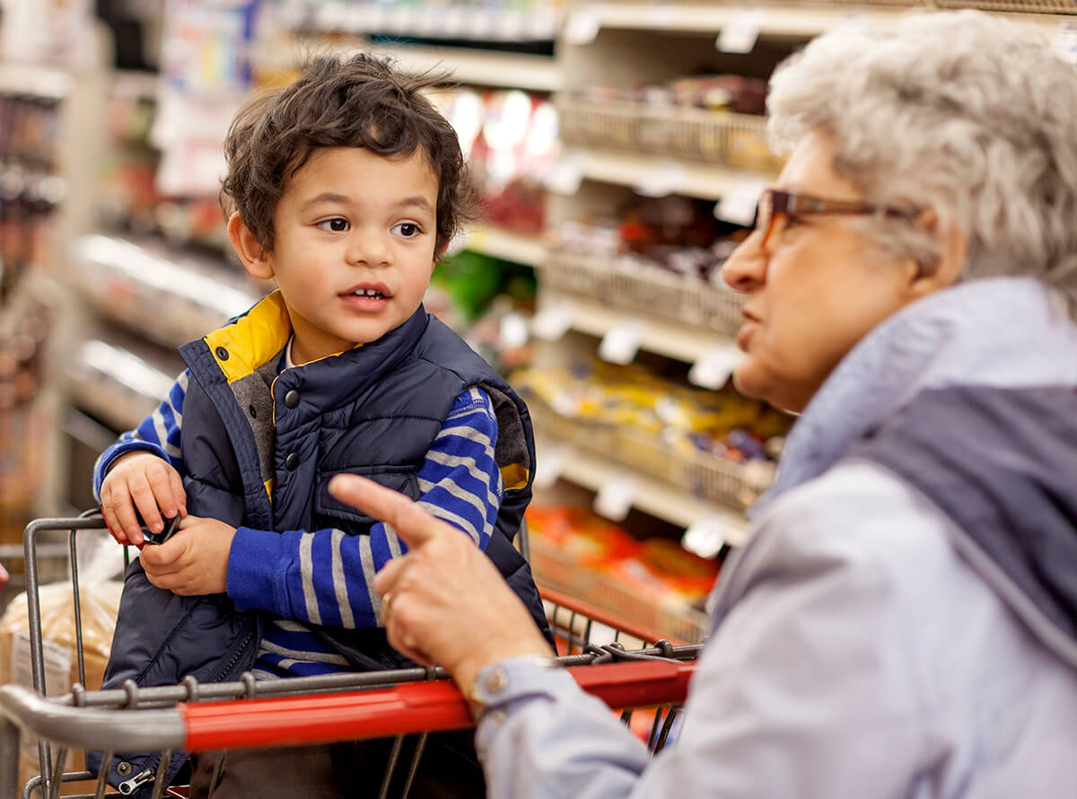 A grandmother talks to her young grandson in a grocery store, pointing to items on the shelf. The young grandson looks at his grandmother from the grocery cart child seat.