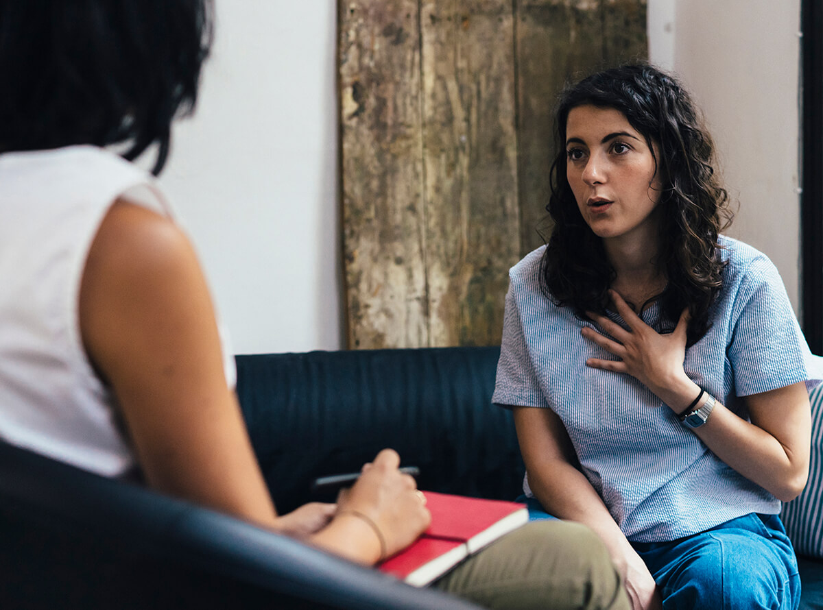 A woman in therapy on a sofa talking to another woman. The first woman has long curly dark hear and wears a blue shirt, the therapist holds a red notebook and pen.