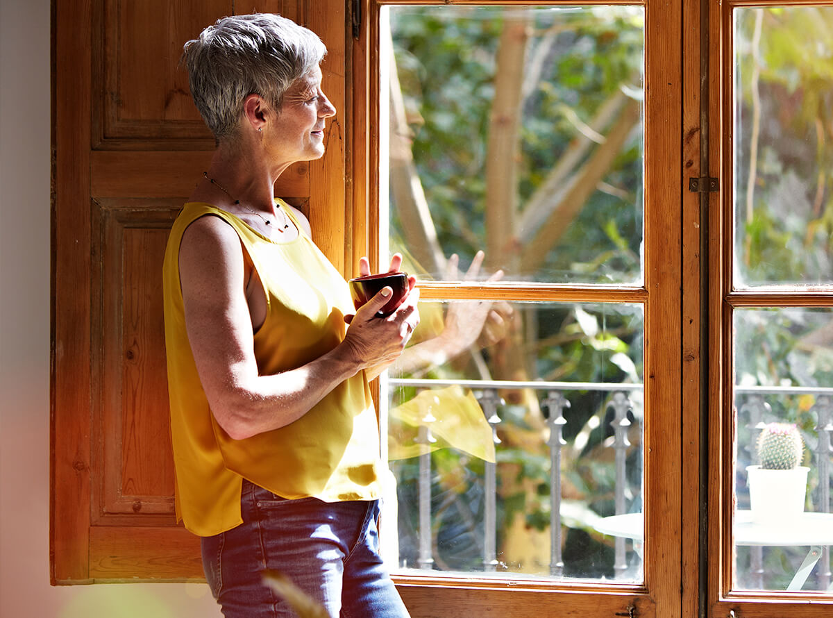 An older woman with short grey hair wearing a yellow shirt smiles holding a cup in her hand while looking out a wood-paned window.