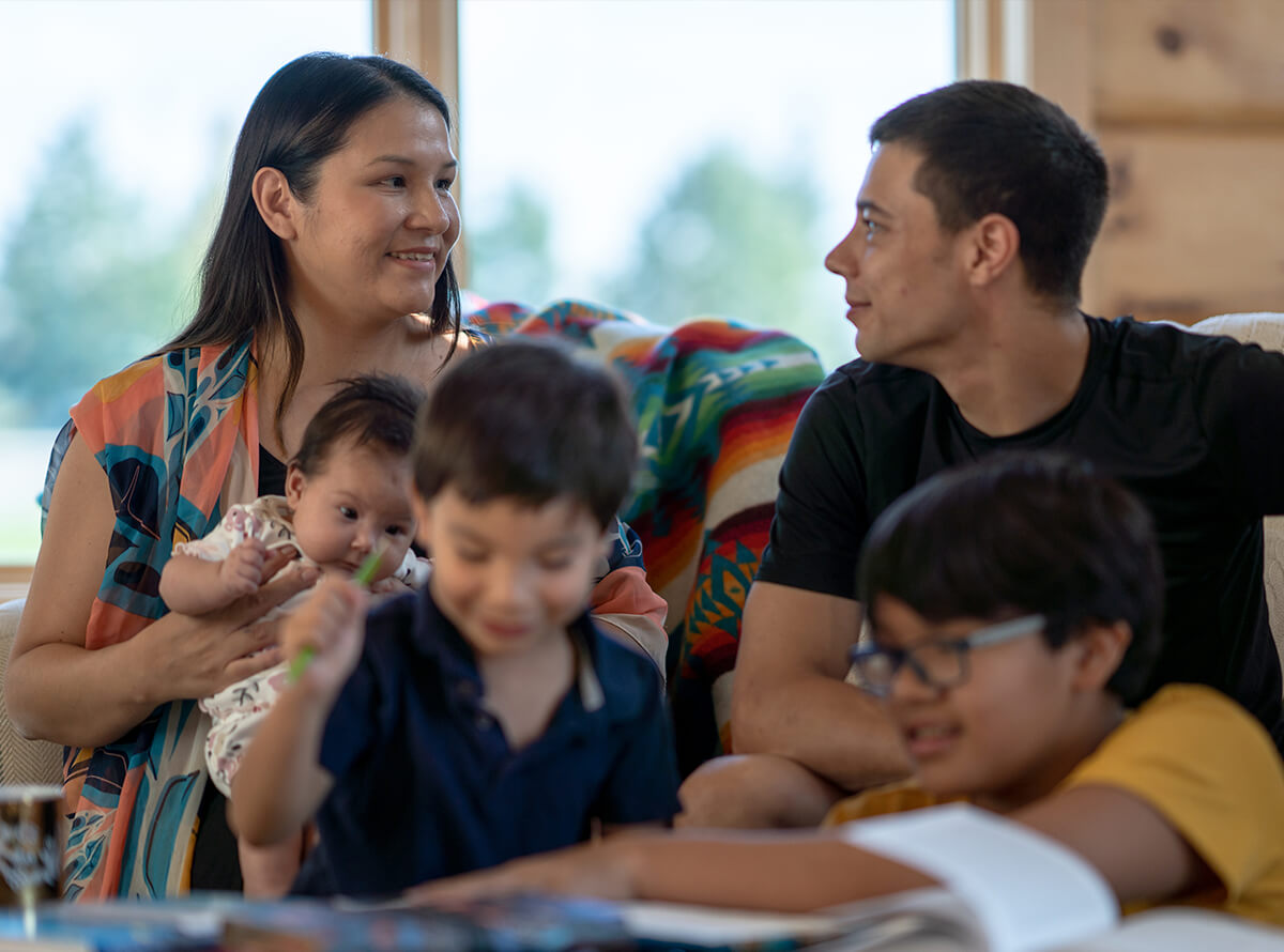 An Indigenous family of five sits together. The woman on the left smiles at her partner while holding a new baby.