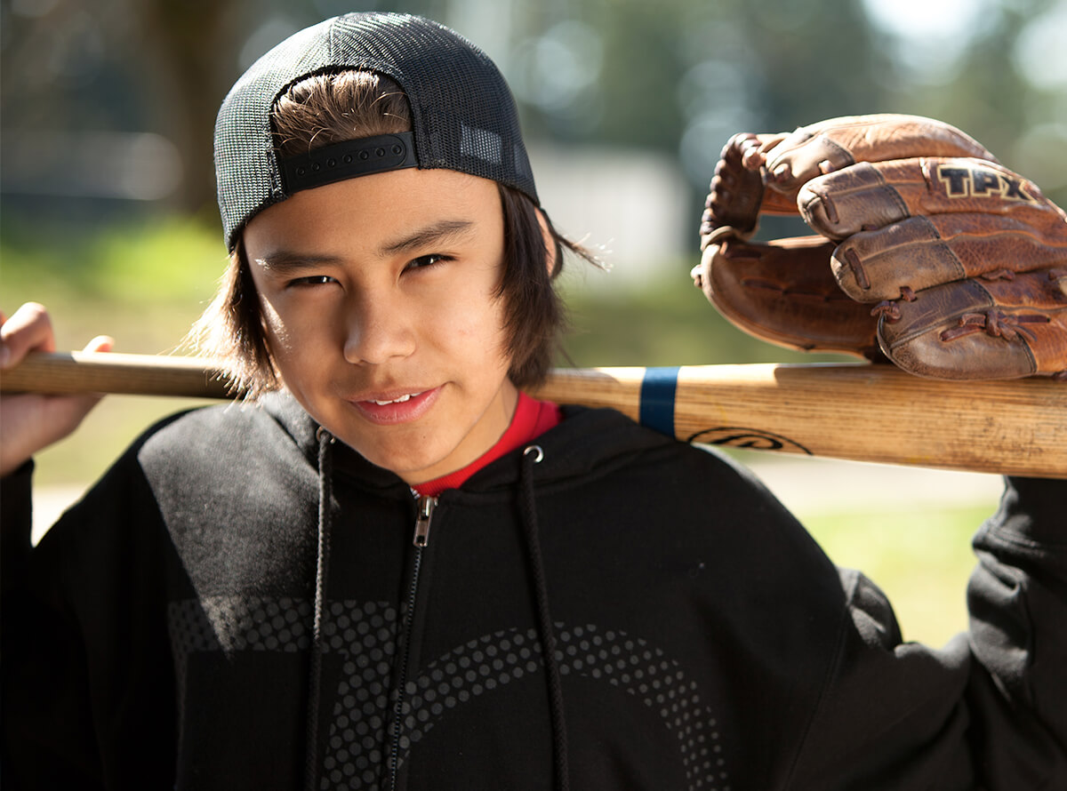 A teenaged Indigenous boy smiles at the camera holding a baseball bat over his shoulders. He wears a baseball glove on his left hand.