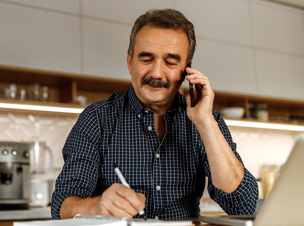 An older man sits in his kitchen while on the phone and writing in a notebook.