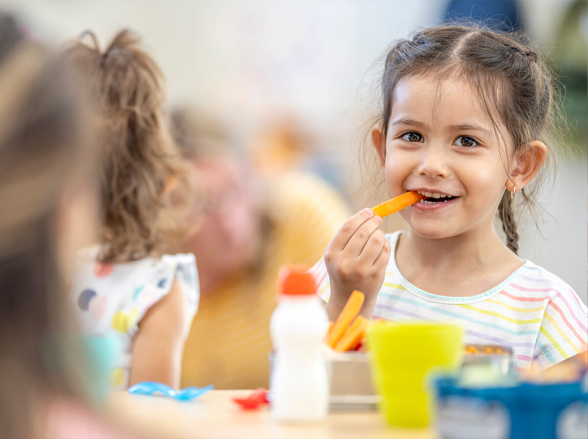 A young girl smiles at the camera while she eats a carrot during lunchtime at school.