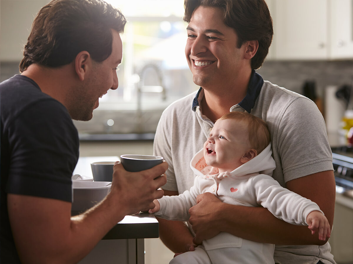 A same-sex couple sit in their kitchen together. One man holds their baby while his partner is making the baby laugh.