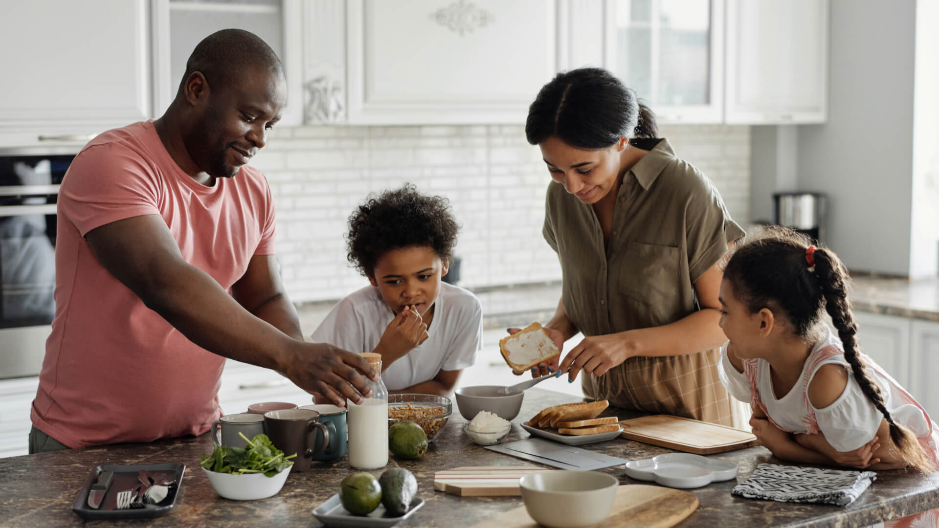 A young Black family stand at their kitchen island while making a snack together, smiling.