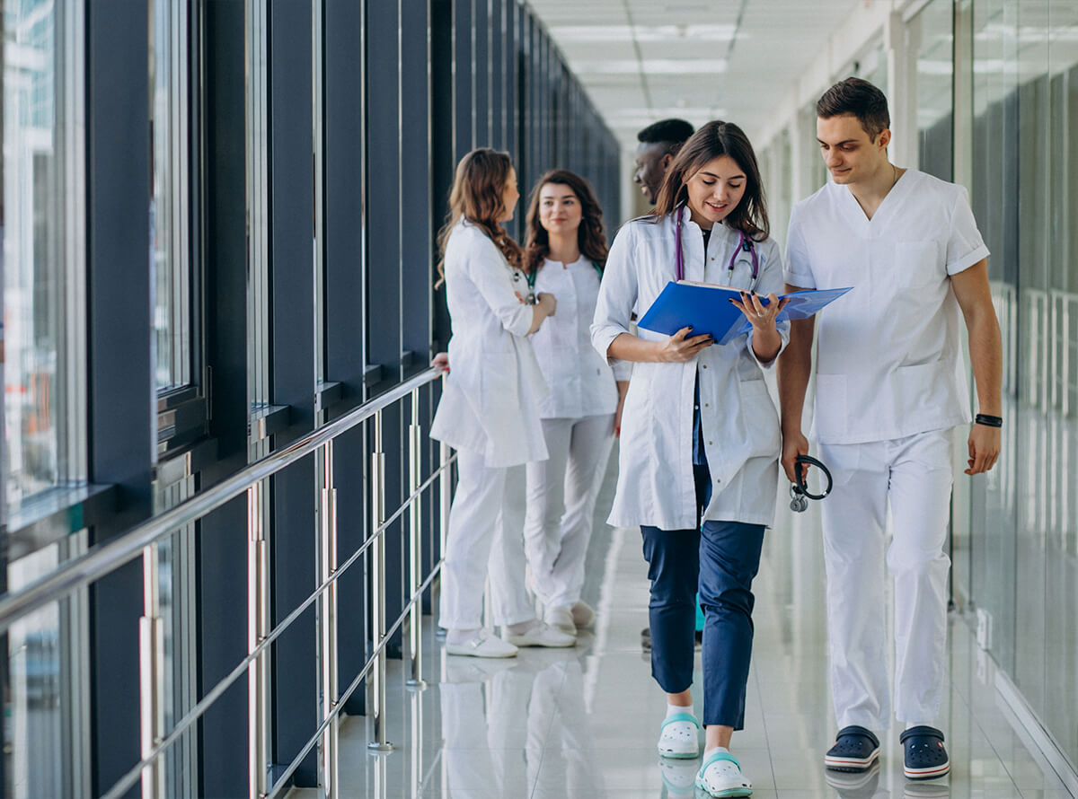 Two young doctors look at a chart while walking down the hall past another group of doctors talking to each other.