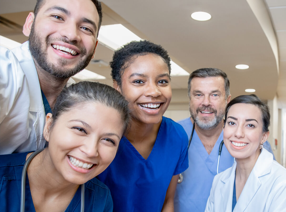 A group of doctors in blue scrubs, two wearing medical jackets, smile at the camera. Four appear to be younger while one is middle-aged.