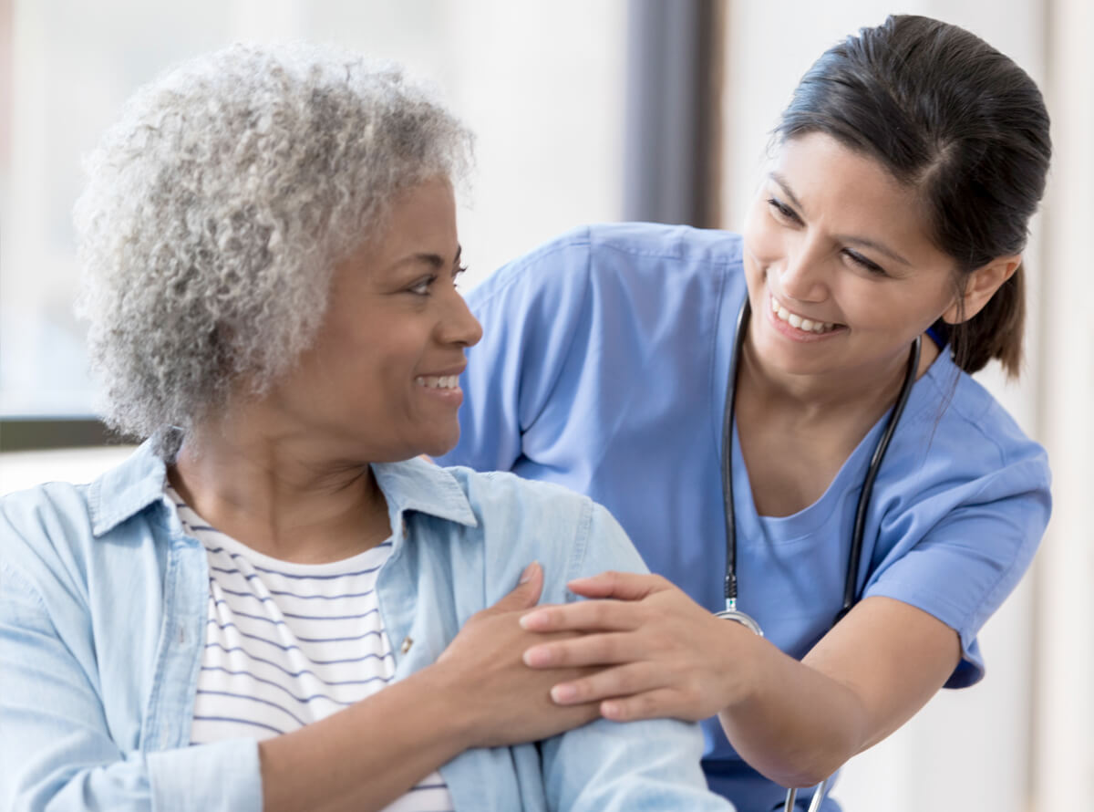 A young nurse stands behind a Black woman who is seated. They are touching hands and smiling at each other.