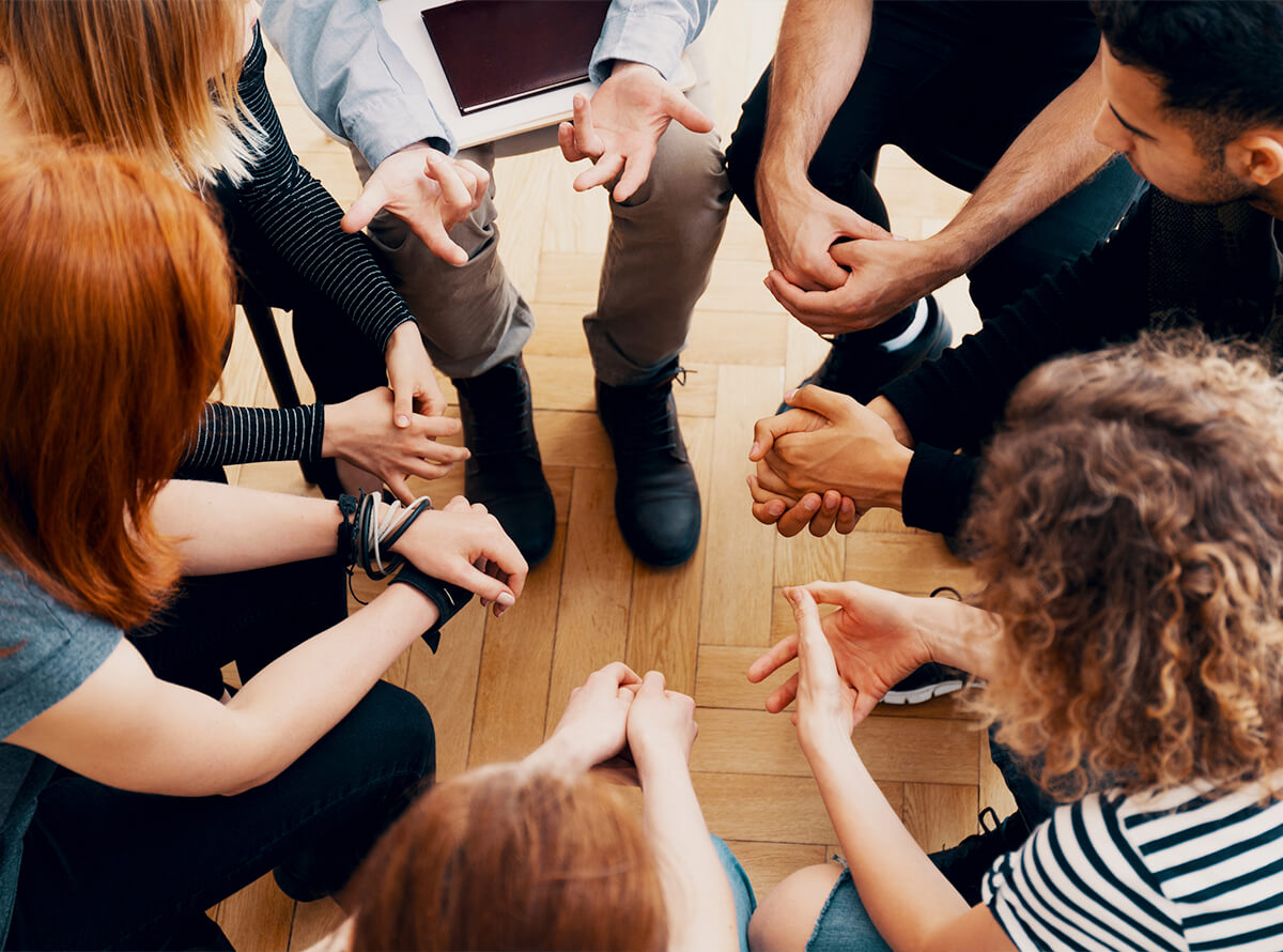 An aerial view of a circle of people sitting in chairs, very close together. We mostly see knees and hands, and the backs of some heads as they all lean in.