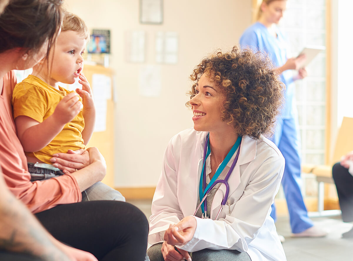 A doctor smiles at her patient, a toddler wearing a yellow shirt who sits in their mother's lap, while a person in scrubs stands in the background.