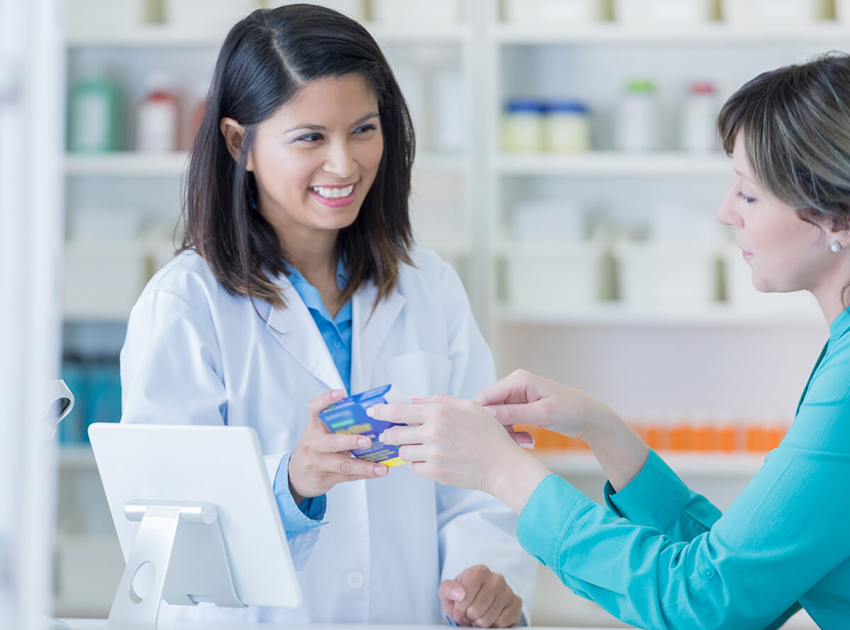 A pharmacist smiles while talking to a woman about a box of medication.