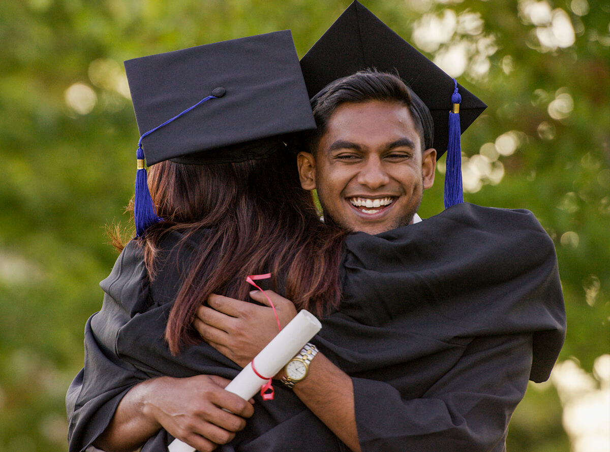 A young man with dark brown skin smiles and hugs his classmate with long red-brown hair. They are wearing graduation robes and hats, and he holds his diploma in one hand.