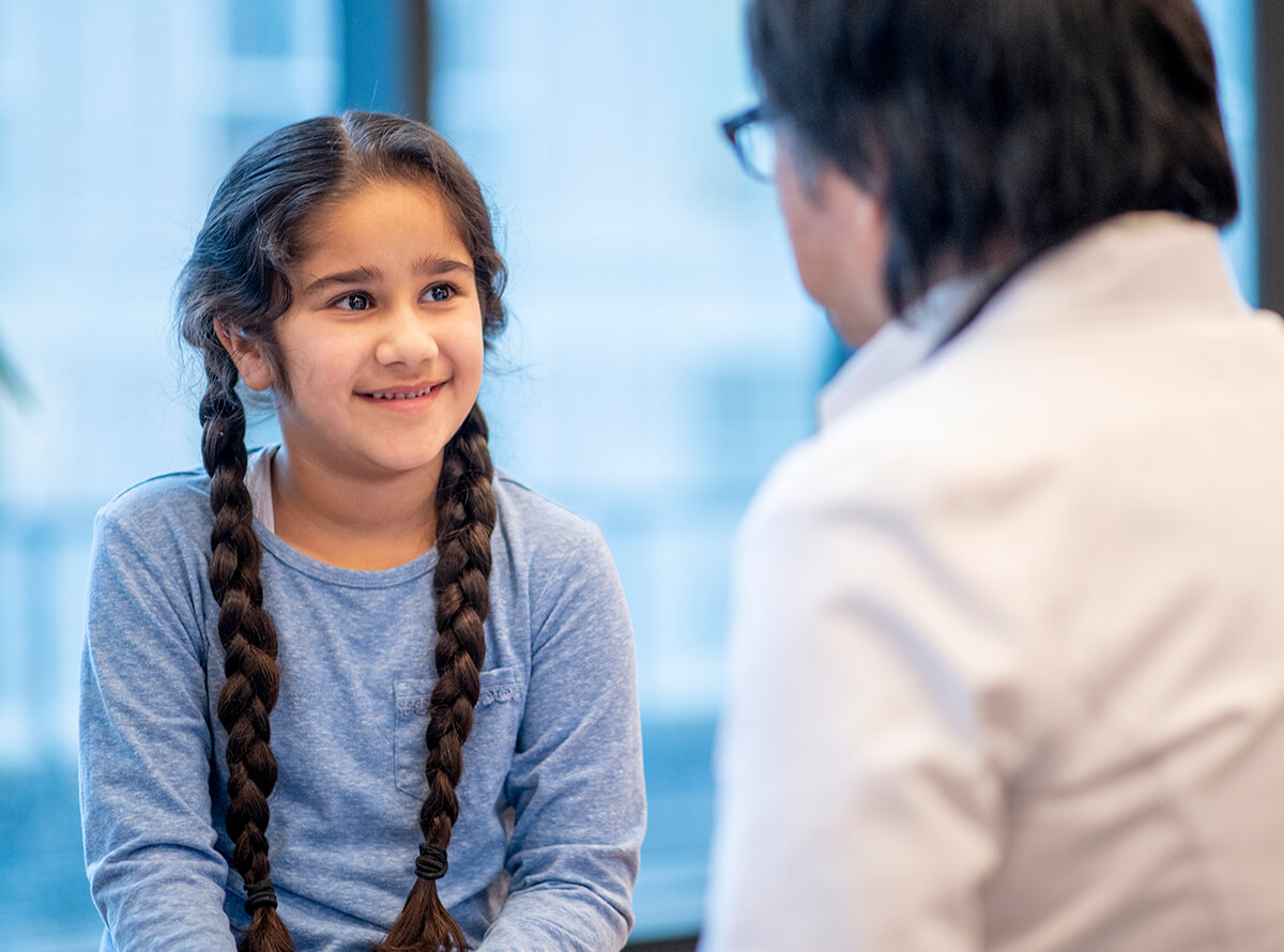 A young girl with long dark braids sits and smiles at a doctor.