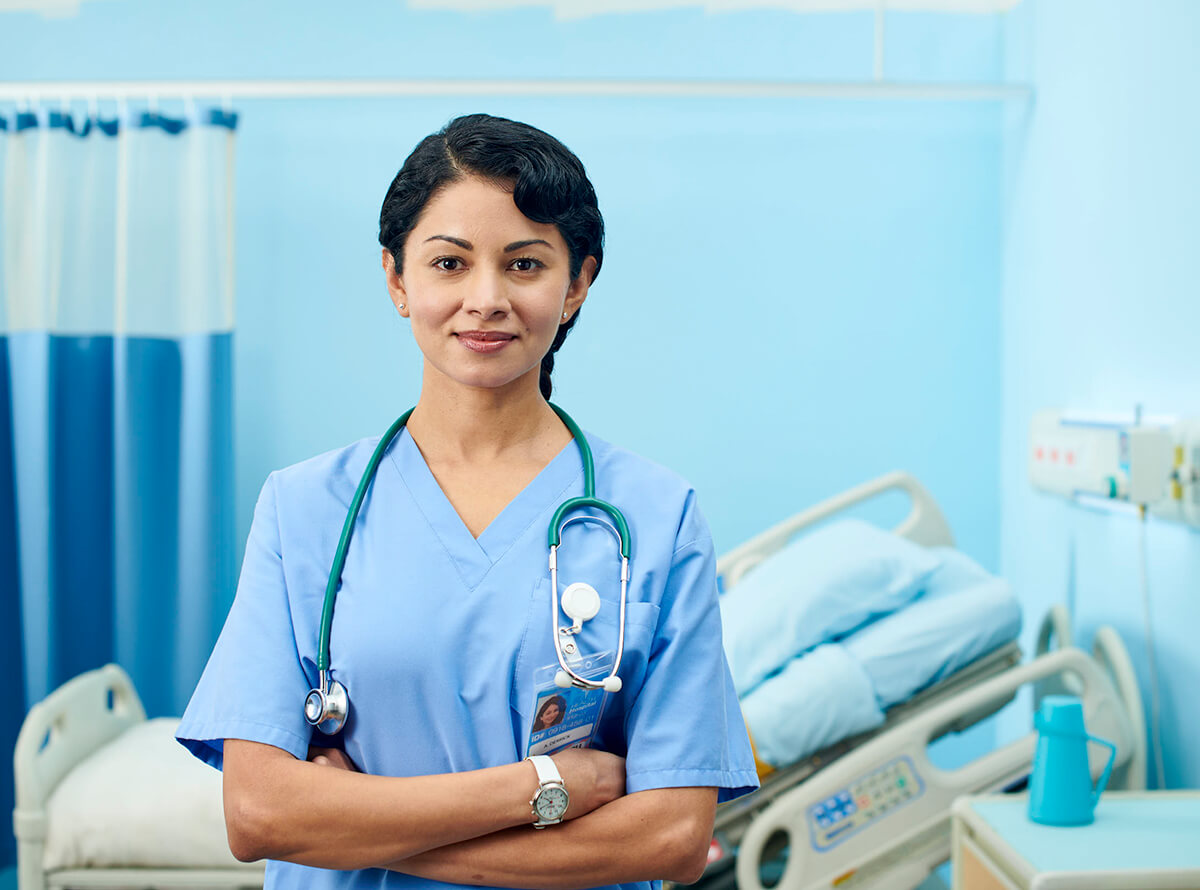 A young woman of colour wears scrubs and a stethoscope while standing in front of a hospital bed in a blue room.
