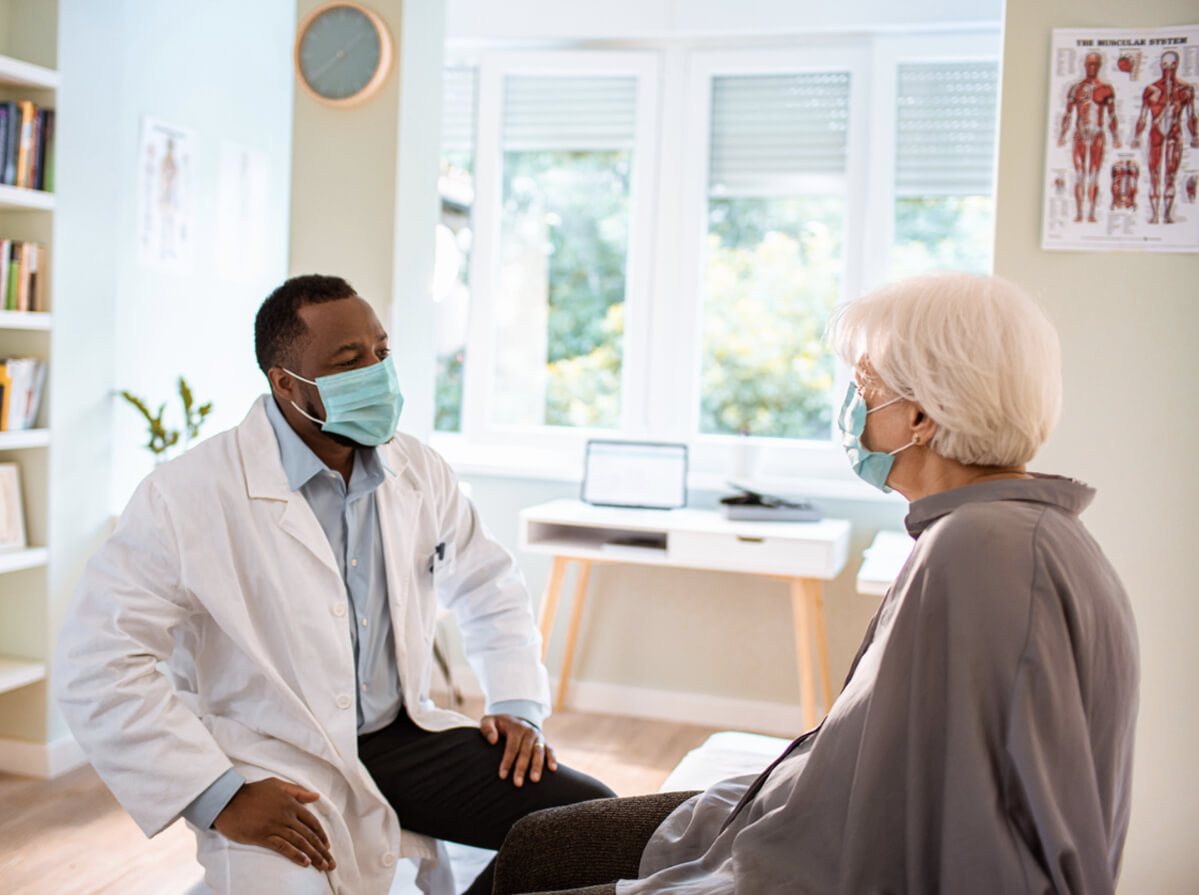 A doctor sits with a patient in a daylight-filled room. They both wear medical masks and appear to be in conversation.