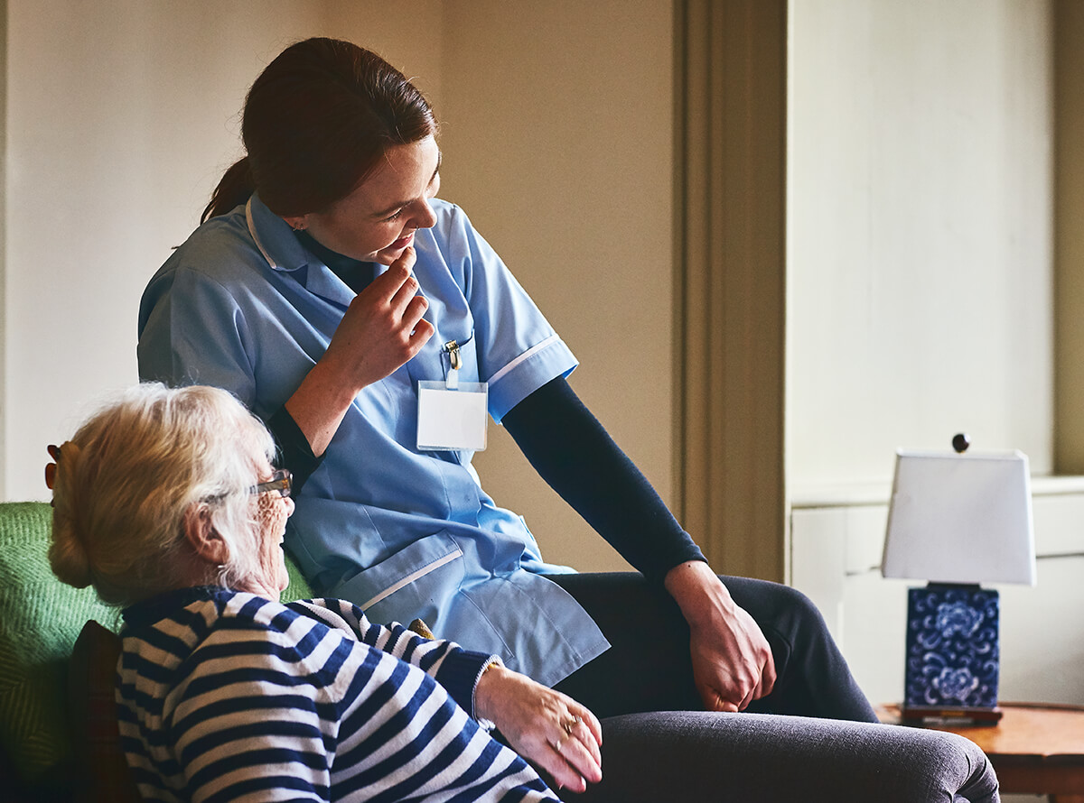 A caretaker and older adult laughing together. The older adult sits in a chair while the caretaker sits on the armrest.