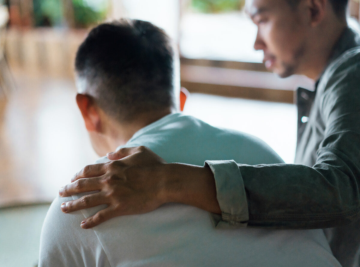 Two men are seen from the back. One sits slumped over with the other man's arm around his shoulders.