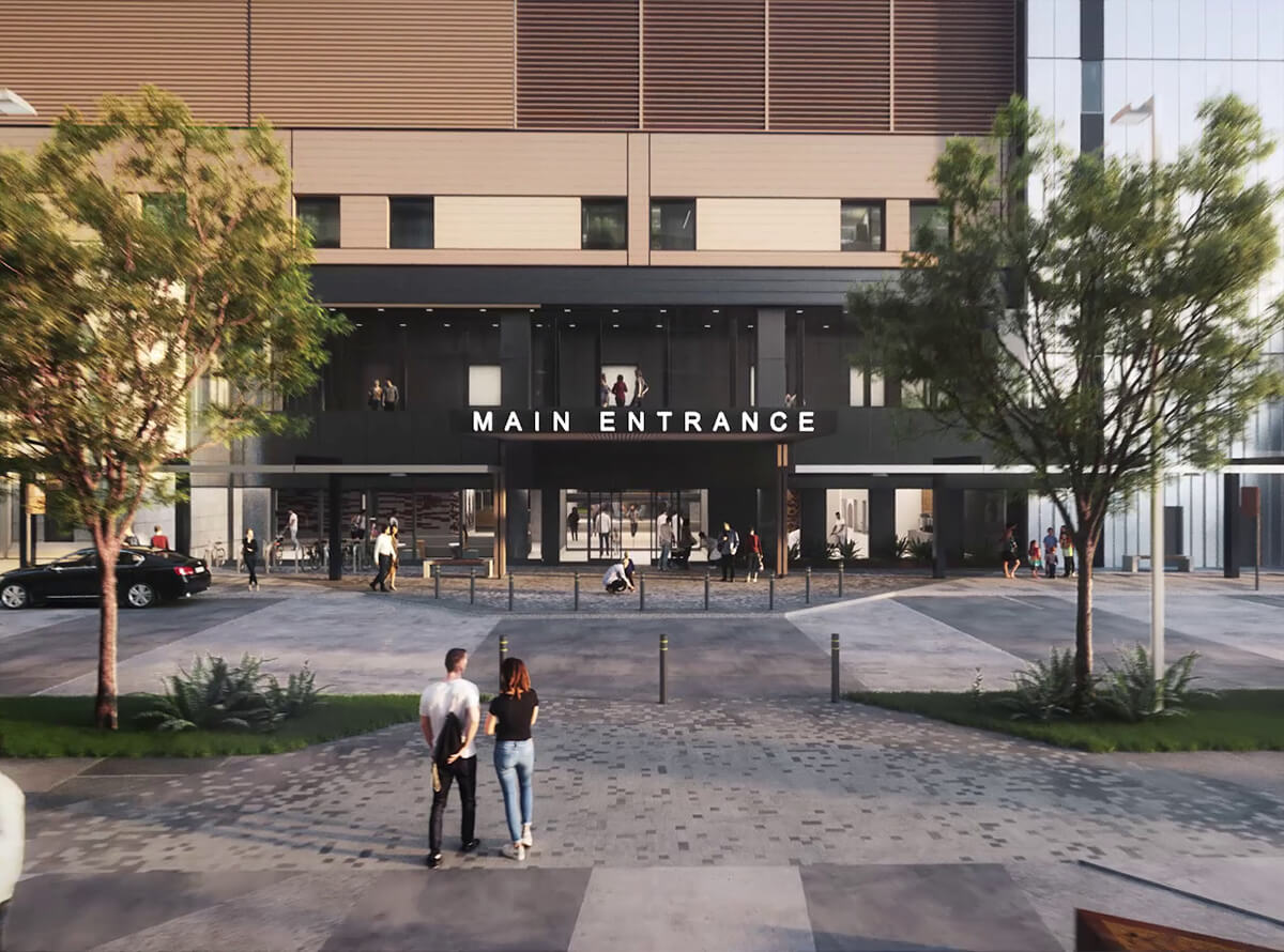 A distance shot of the Main Entrance of a building with trees on pavilions in front. Above the doors are the 3D words "Main Entrance."