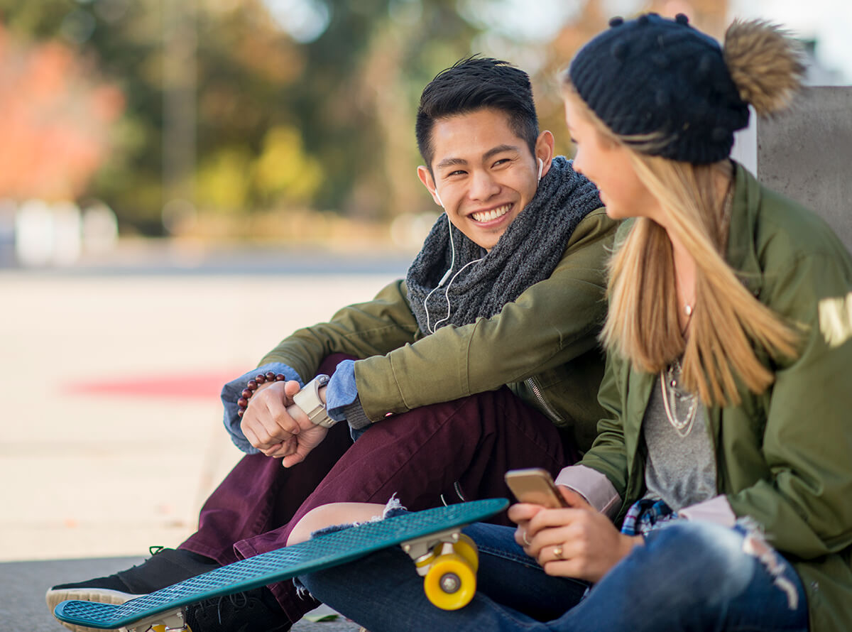 Two youth are seen sitting outside at a skate park wearing green jackets and smiling at each other. The woman on the right holds a phone and has a penny board propped up on her knee.