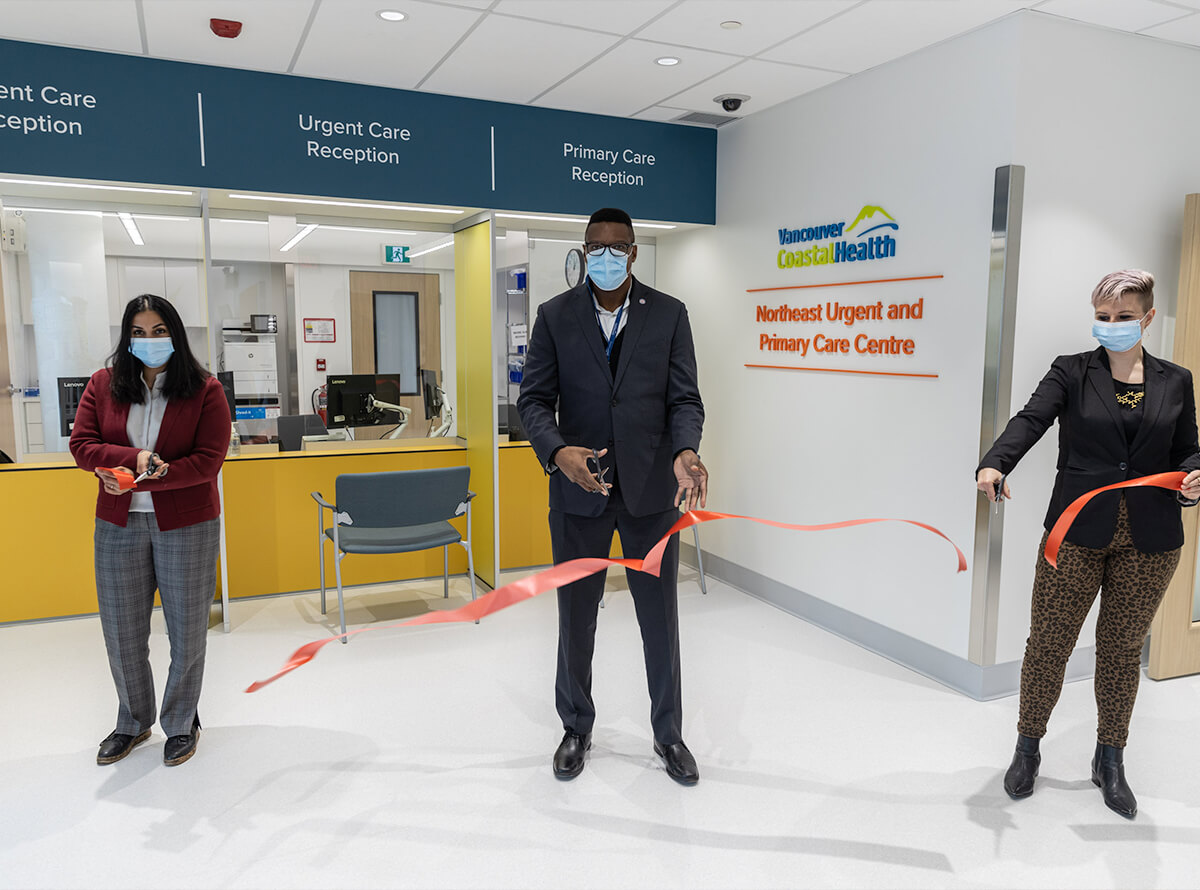 Hospital executive staff are seen cutting a ribbon in the reception area of a Vancouver Coastal Health hospital. the wall reads "Northeast Urgent and Primary Care Centre".