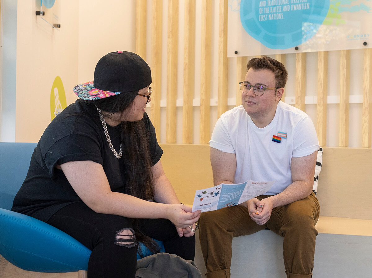 Two young adults sit and talk holding a flyer. One wears all black with ripped jeans, a silver chain, and backwards cap. The other is a masculine-presenting person wearing a white t-shirt and pins of the transgender and 2SLGBTQ+ flags.