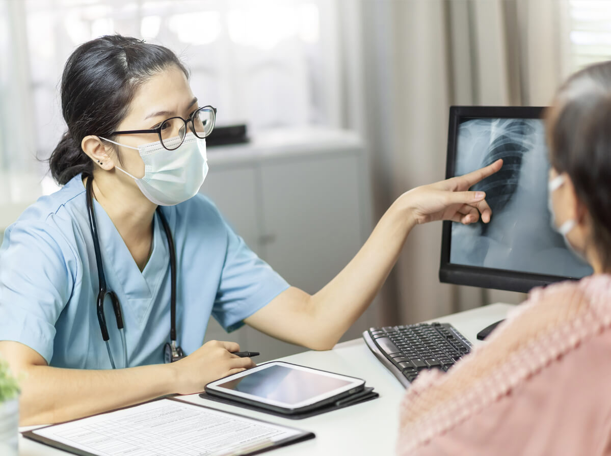 An Asian woman in scrubs wearing a stethoscope points to an Xray while talking to a patient. They are both wearing medical masks.