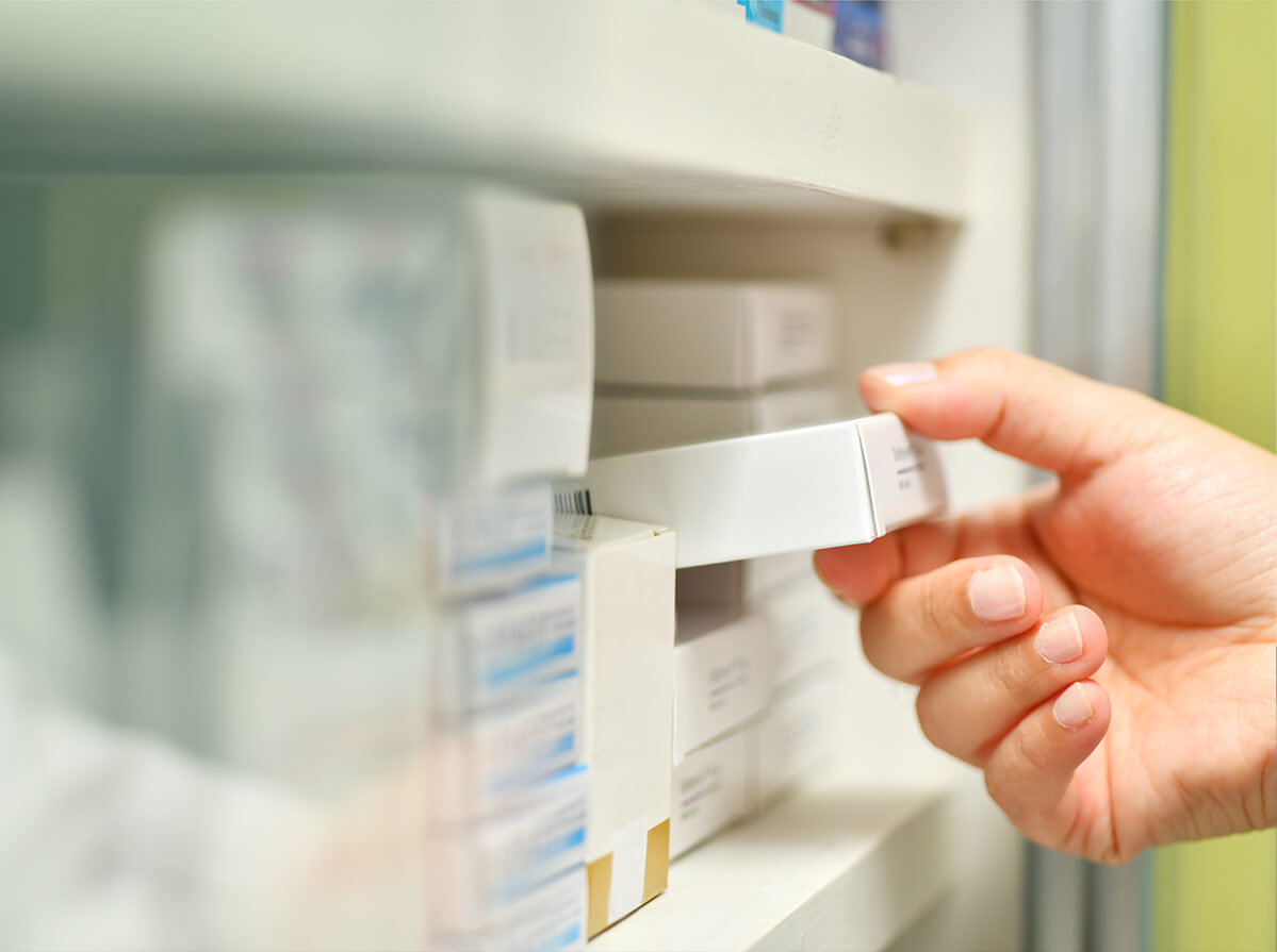 The hand of a pharmacist is seen pulling a box of medication from the shelf.