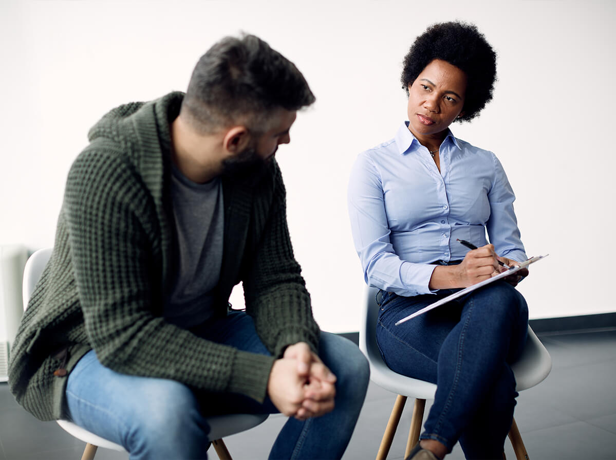 Two people sit next to each other in white chairs against a white wall background. The man on the left is slouching while looking at the woman on the right who holds a clipboard.