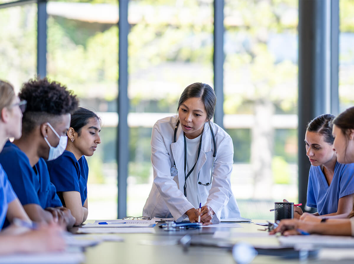 A doctor in a med jacket stands at the end of a table talking to a group of young doctors in blue scrubs.