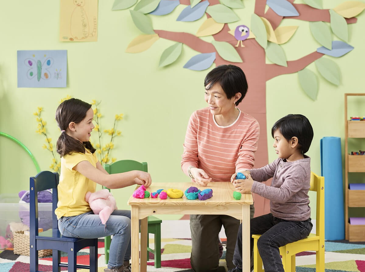 A childcare worker sitting at a small table with two young children. The worker and one child are smiling while paying attention to the other child while they all play with Play-Doh.