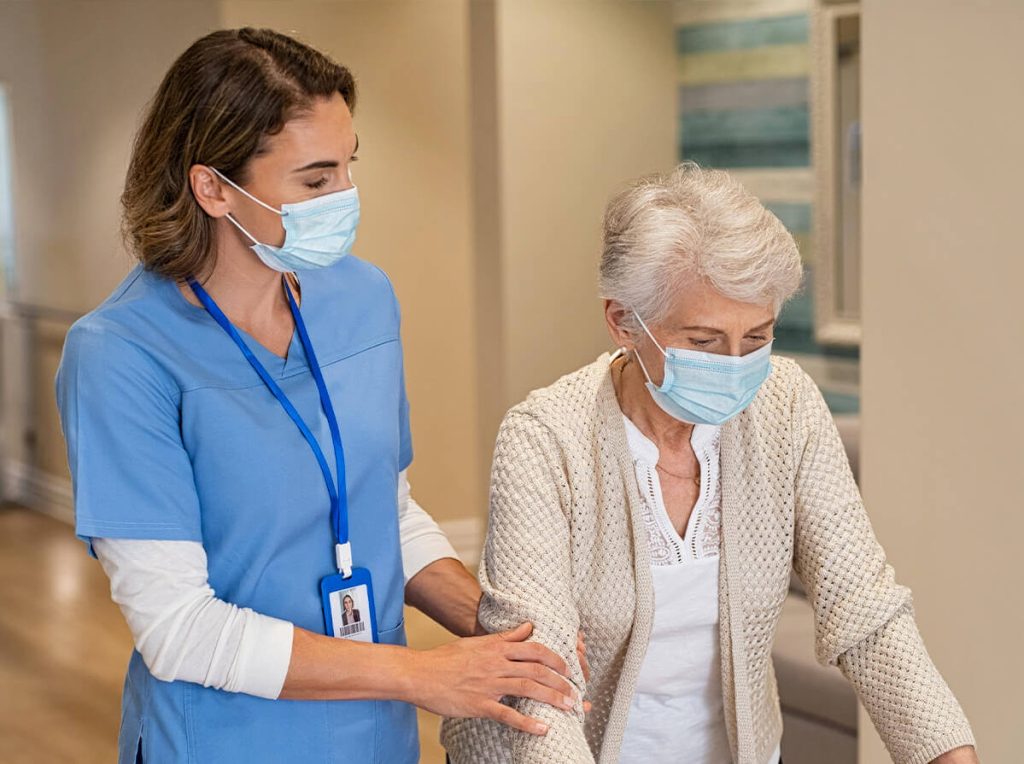 A caretaker in blue scrubs holds the arm of an older adult in a care facility. They are both wearing medical masks and the patient is seen wearing a cardigan, presumably using a walker.