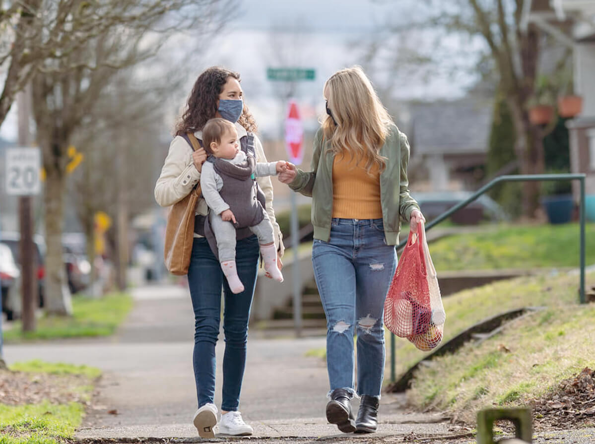 Two woman walk together on a city sidewalk carrying reusable grocery bags. One woman is carrying her baby while the other holds the baby's hand. They are both wearing reusable cloth masks.