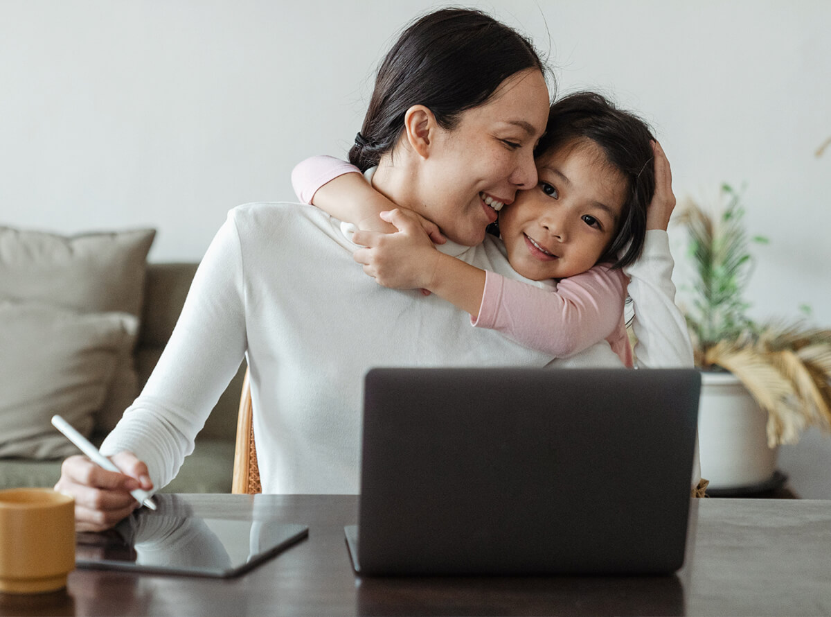 An Asian woman sits at her desk at home while her daughter puts her arms around her and her head on her mother's shoulder. The daughter is smiling at the camera while the woman smiles at her child.