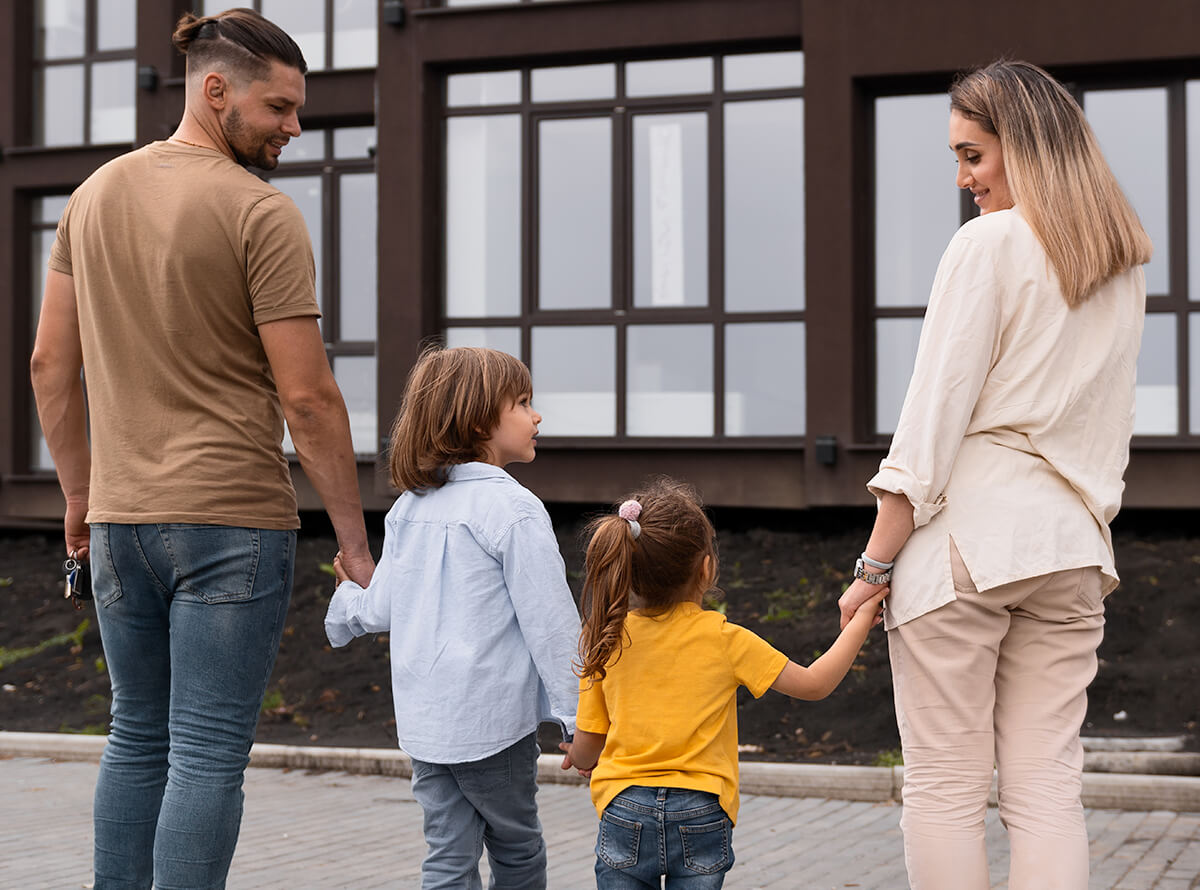 A couple hold their two children's hands in front of a building complex, smiling at the children.