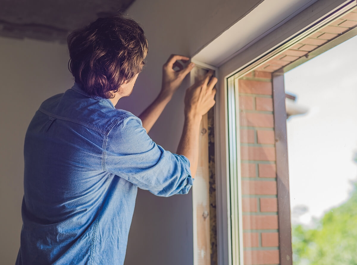 A white man is seen from behind while he works on maintaining a window sill's drywall inside a room.