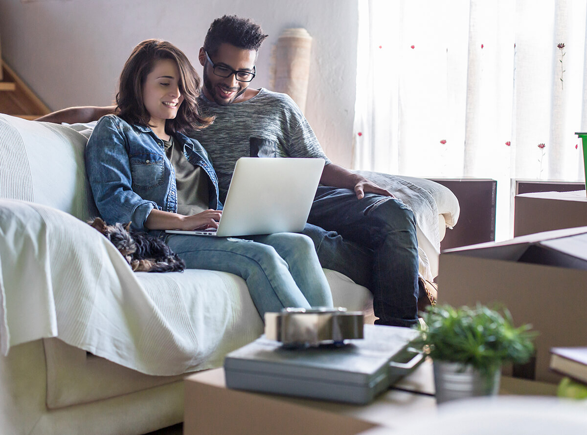 A young biracial couple sit on a sofa together looking at a laptop. Moving boxes appear around them.