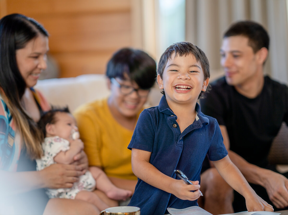 A young boy smiles wide at the camera standing in front of his family members. Three adults are seen in the background, with the woman on the left holding a young baby in her lap.