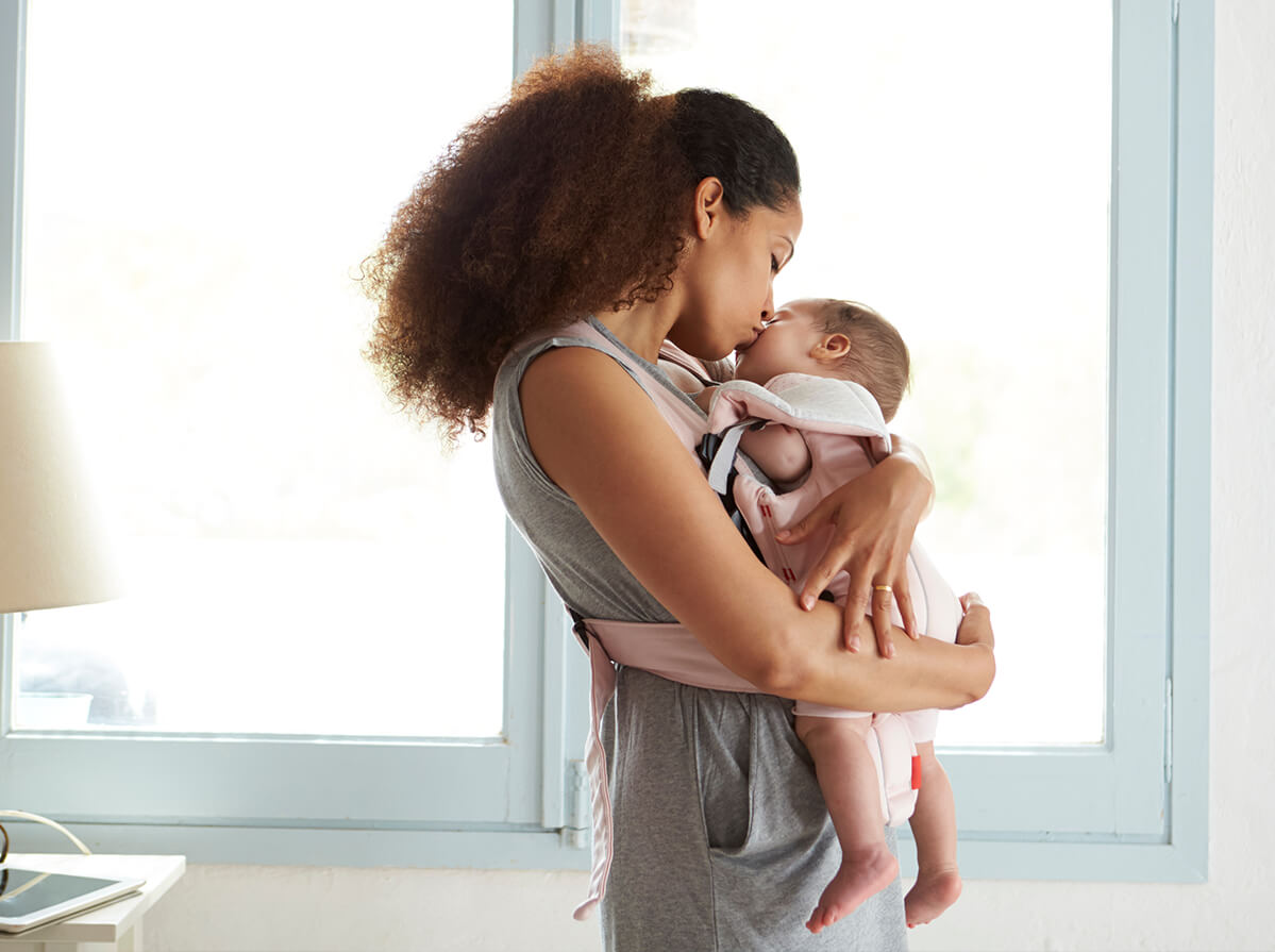 A woman is wearing her baby, has her arms wrapped around the infant and kisses their cheek. They are in front of a window with light blue trim.